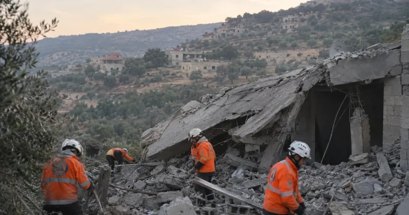 Rescue workers in orange high-visibility jackets and white helmets search the rubble of a collapsed concrete house on a hillside in southern Lebanon, with olive terraces and village homes behind them at dawn