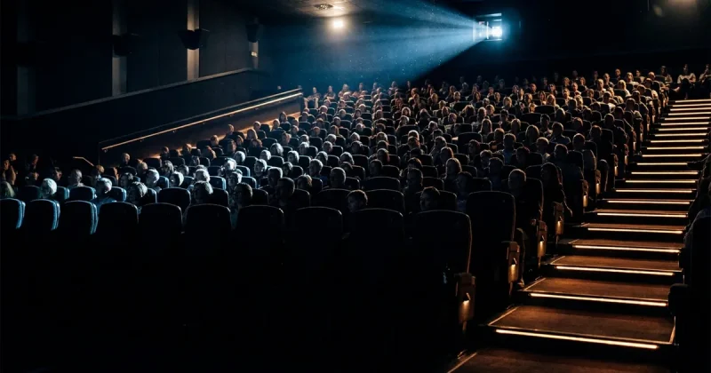 A completely full multiplex auditorium seen from the front, audience faces lit by the glow of an off-frame screen with the projector beam cutting through the dark
