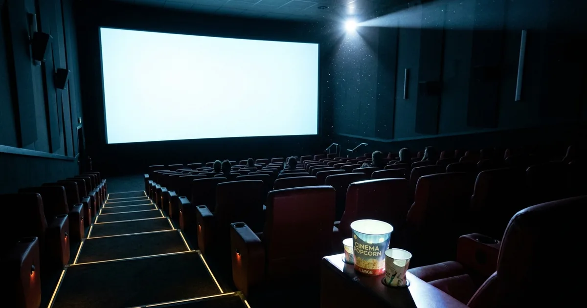 A mostly empty cinema auditorium seen from the back row, rows of dark red recliners facing a bright blank screen with a popcorn tub on a foreground armrest