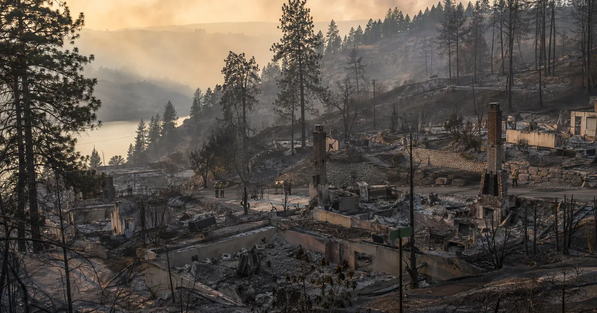 Burned homes reduced to concrete foundations with brick chimneys and a street sign still standing on a scorched pine bluff above the Spokane River, smoke haze in the valley and firefighters in the distance