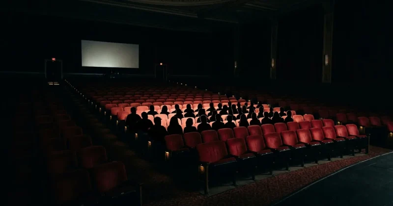 A darkened cinema auditorium where a small cluster of center rows is filled with silhouetted moviegoers while hundreds of red seats around them sit empty