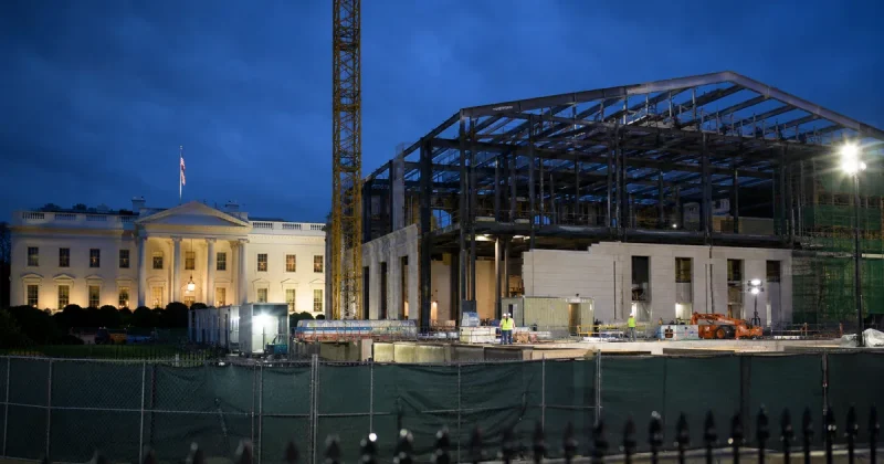 The White House lit at dusk beside a floodlit construction site where the East Wing stood, showing an exposed steel frame, a tower crane and workers behind green site fencing