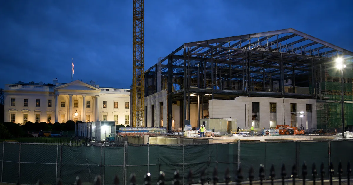 The White House lit at dusk beside a floodlit construction site where the East Wing stood, showing an exposed steel frame, a tower crane and workers behind green site fencing