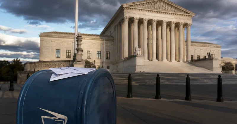 A blue United States Postal Service collection mailbox with white ballot envelopes resting on top stands on the sidewalk in front of the U.S. Supreme Court building in Washington under a heavy overcast sky.