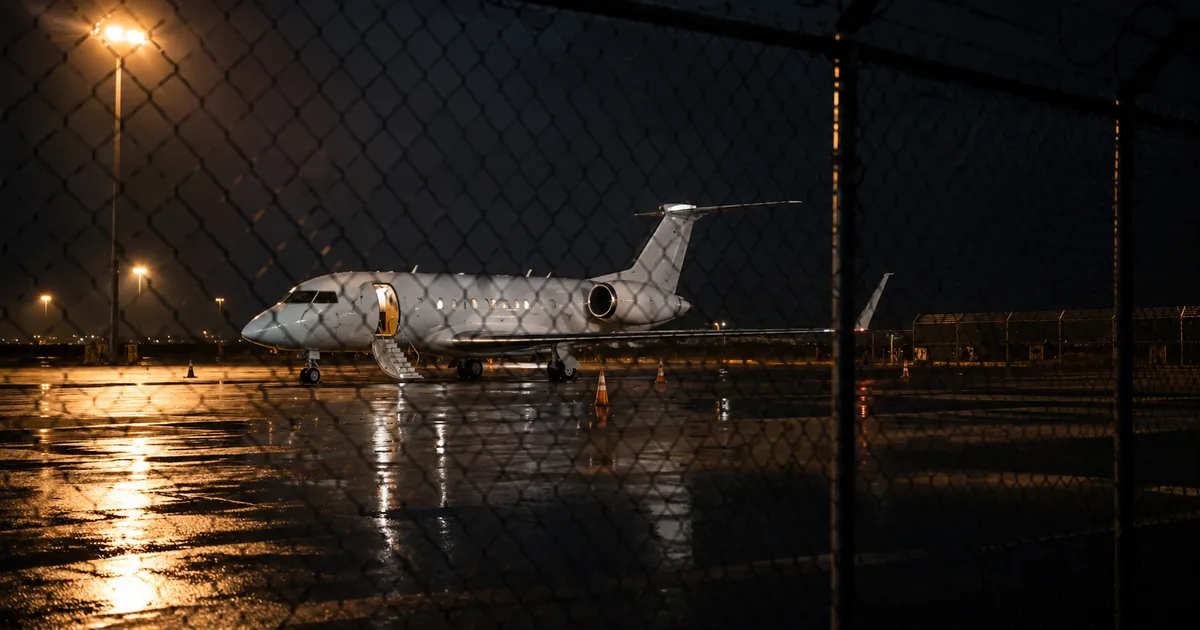 An unmarked white charter jet with its airstair lowered sits on a rain-slicked apron at night, seen through a chain-link perimeter fence under sodium floodlights