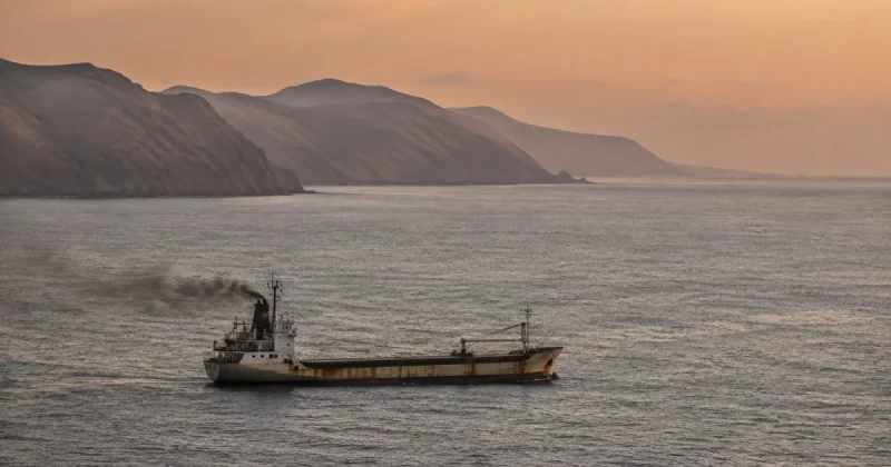 A small rust-streaked general cargo coaster sails alone through an empty strait at dawn, with an arid mountainous coastline in the background
