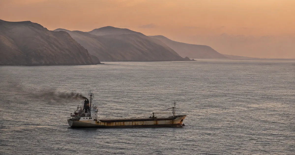 A small rust-streaked general cargo coaster sails alone through an empty strait at dawn, with an arid mountainous coastline in the background