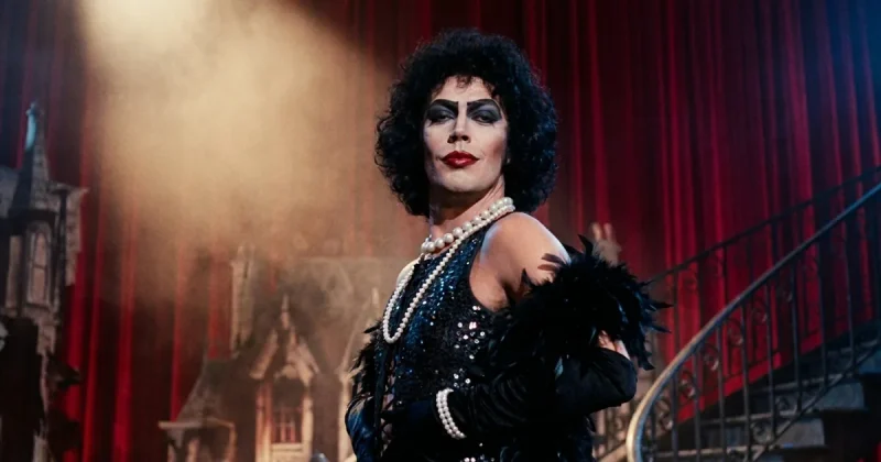 Tim Curry in character as Dr. Frank-N-Furter, in teased black curls, heavy stage makeup, red lipstick, pearls and a black sequinned costume with a feather boa, lit by a follow-spot on a red-curtained stage