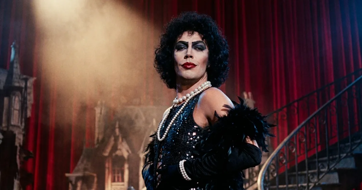 Tim Curry in character as Dr. Frank-N-Furter, in teased black curls, heavy stage makeup, red lipstick, pearls and a black sequinned costume with a feather boa, lit by a follow-spot on a red-curtained stage
