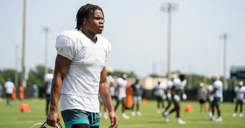 Football player in a plain white practice jersey and teal shorts carries his helmet at his side on a training camp field, teammates blurred behind him in the summer heat
