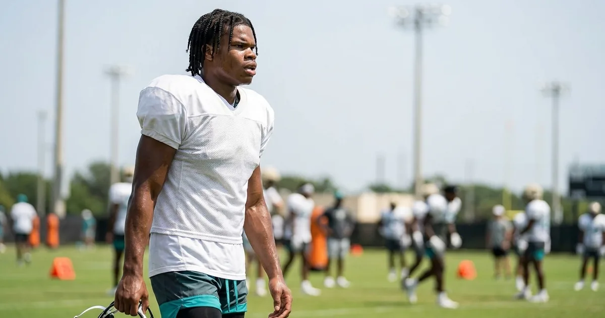 Football player in a plain white practice jersey and teal shorts carries his helmet at his side on a training camp field, teammates blurred behind him in the summer heat