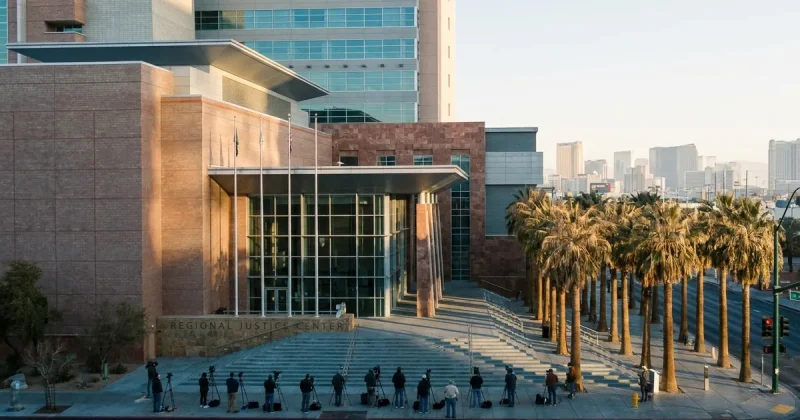 News camera operators set up on the steps of the Las Vegas Regional Justice Center in early morning light with palm trees and the city skyline behind