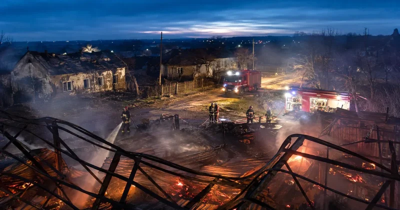 Firefighters hose down the collapsed, smouldering steel frame of a burned-out warehouse at night while two fire engines stand on a village road beside scorched single-storey houses