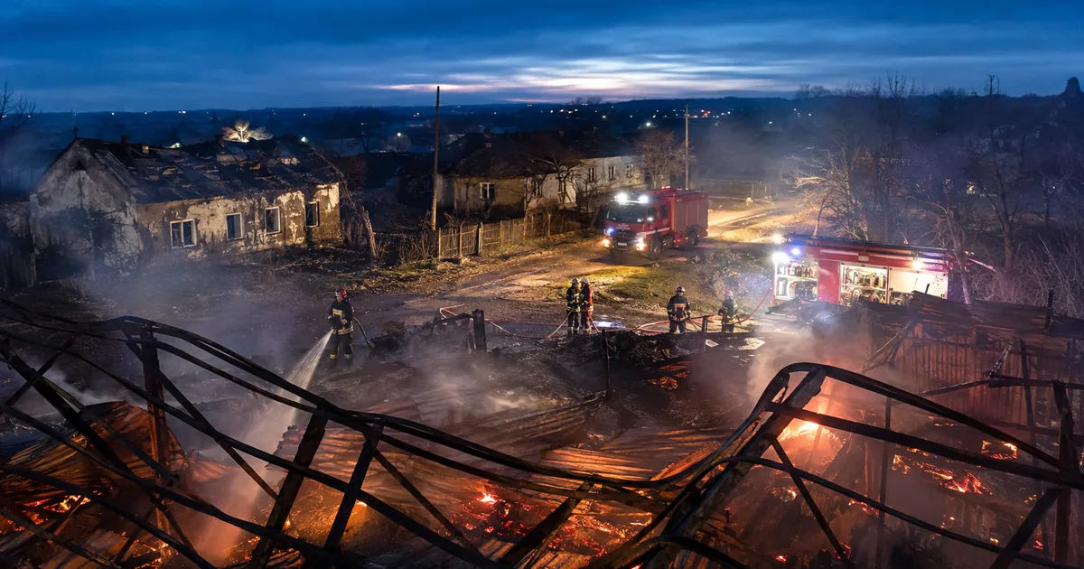 Firefighters hose down the collapsed, smouldering steel frame of a burned-out warehouse at night while two fire engines stand on a village road beside scorched single-storey houses