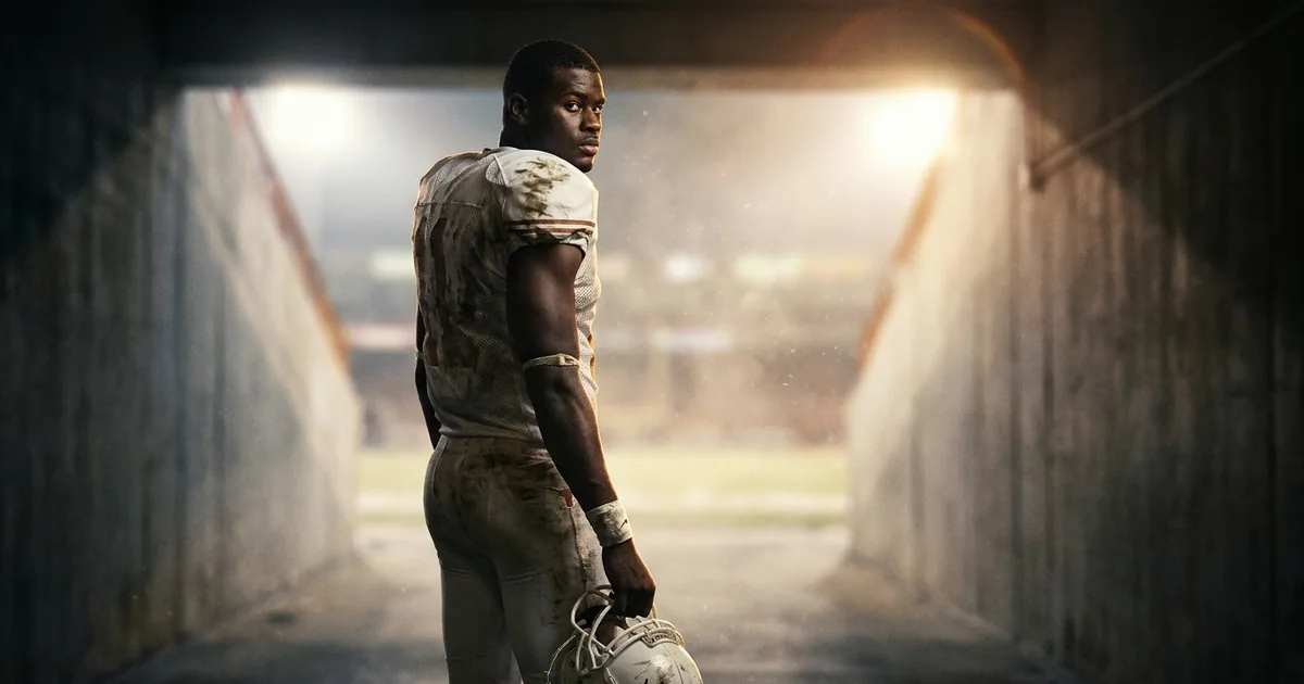A young football quarterback in a mud-streaked white and burnt-orange uniform holds his helmet and looks back over his shoulder from a dark stadium tunnel toward the floodlit field