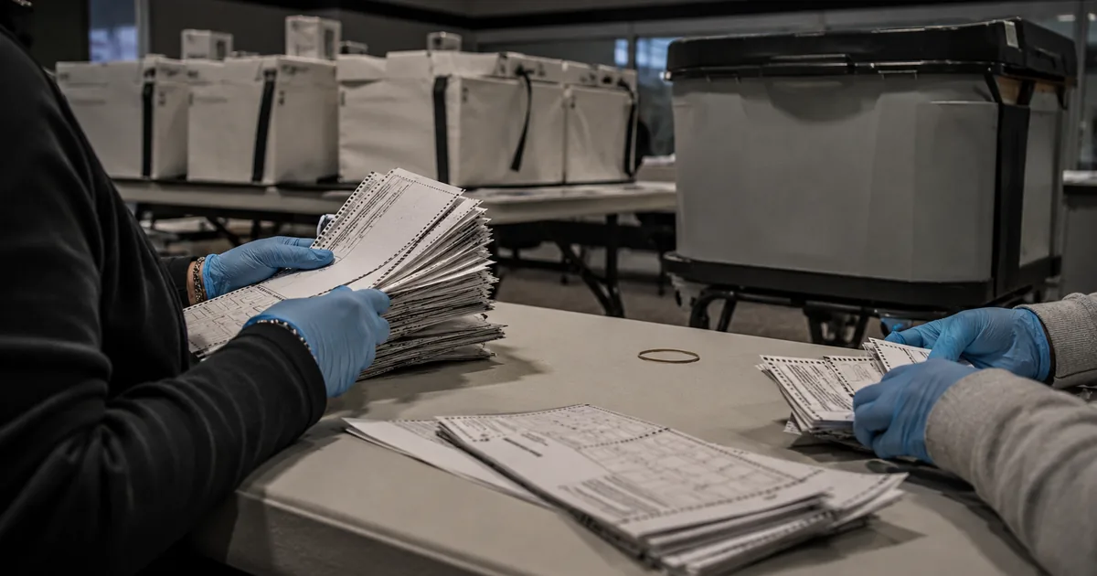 Two pairs of blue-gloved hands sorting stacks of paper ballots on a table beside sealed ballot transfer boxes in an election office