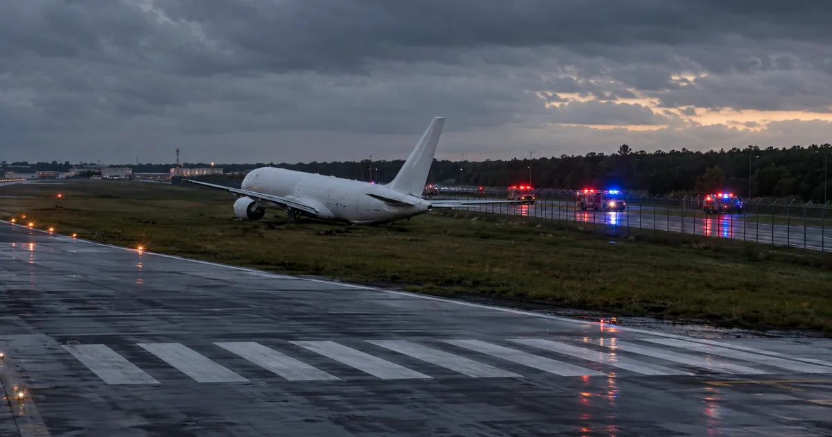 A white widebody cargo freighter stopped on grass past the end of an airport runway at dusk, with a perimeter fence and public road beyond and emergency lights in the distance