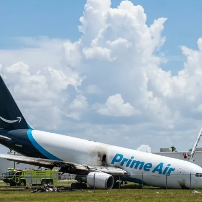 An Amazon Prime Air Boeing 767 freighter rests nose-down on grass past the Miami airport fence with a scorched wing, surrounded by lime-green fire rescue trucks and a fuel tanker under a bright cloudy sky