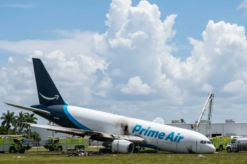 An Amazon Prime Air Boeing 767 freighter rests nose-down on grass past the Miami airport fence with a scorched wing, surrounded by lime-green fire rescue trucks and a fuel tanker under a bright cloudy sky