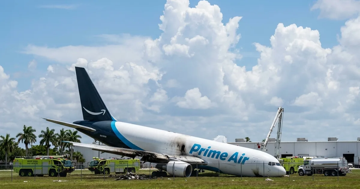 An Amazon Prime Air Boeing 767 freighter rests nose-down on grass past the Miami airport fence with a scorched wing, surrounded by lime-green fire rescue trucks and a fuel tanker under a bright cloudy sky