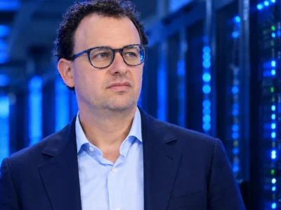 Anthropic CEO Dario Amodei, in glasses and a navy blazer, standing in a dim server hall lit by blue data center racks