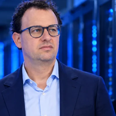 Anthropic CEO Dario Amodei, in glasses and a navy blazer, standing in a dim server hall lit by blue data center racks