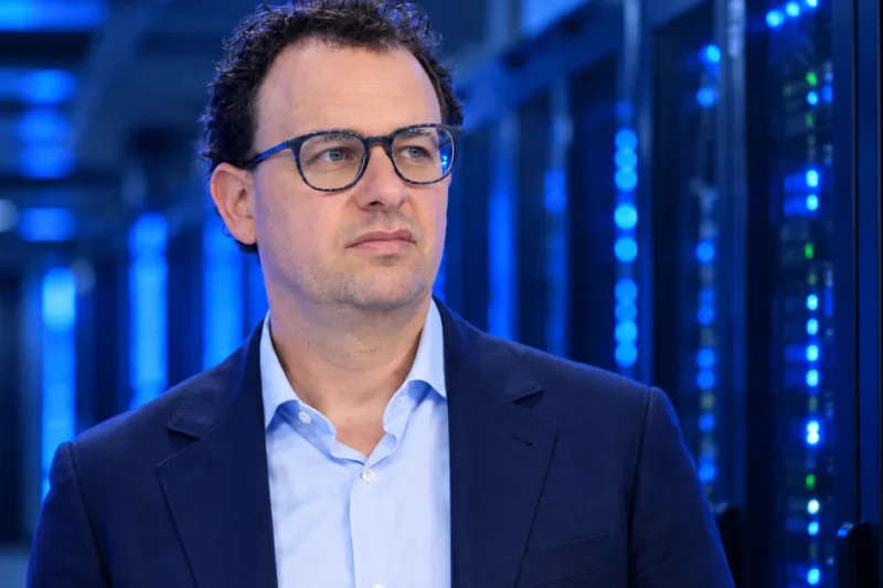 Anthropic CEO Dario Amodei, in glasses and a navy blazer, standing in a dim server hall lit by blue data center racks