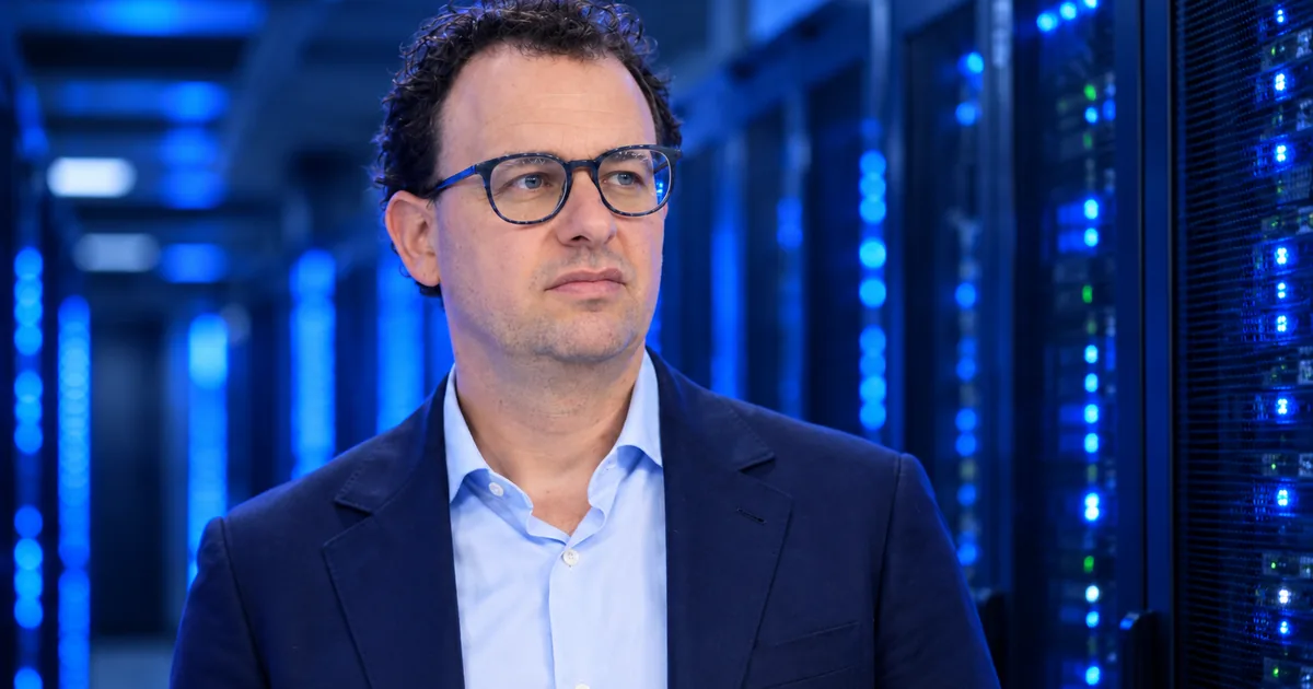 Anthropic CEO Dario Amodei, in glasses and a navy blazer, standing in a dim server hall lit by blue data center racks