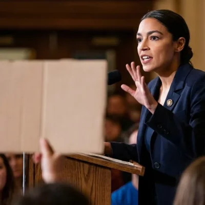 Alexandria Ocasio-Cortez in a navy blazer speaks at a wooden podium with one hand raised as supporters hold up blank placards in the foreground