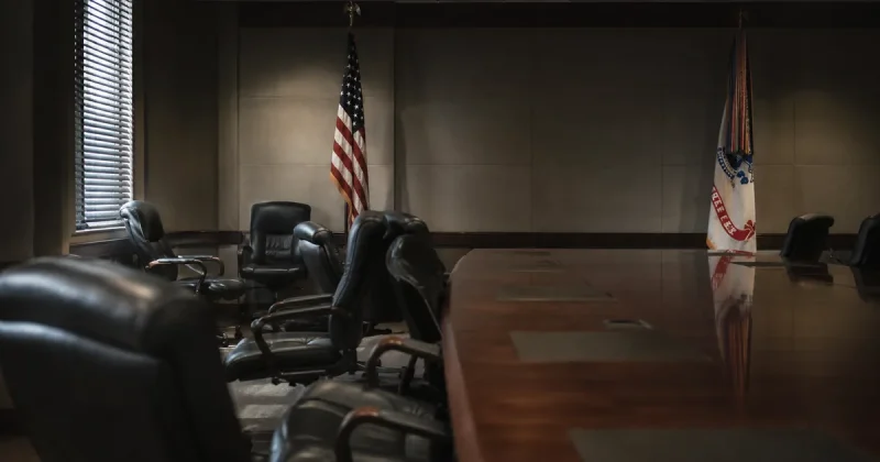A Pentagon briefing room with several empty leather chairs pushed back from a long conference table, US Army and American flags furled against the wall