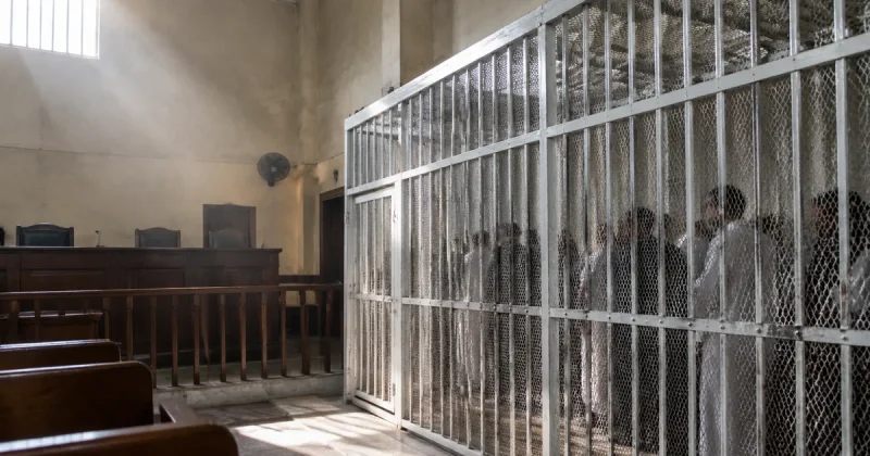 A tall white steel defendants cage stands in an Egyptian criminal courtroom, lit by daylight from a high window, with an empty judges bench to one side