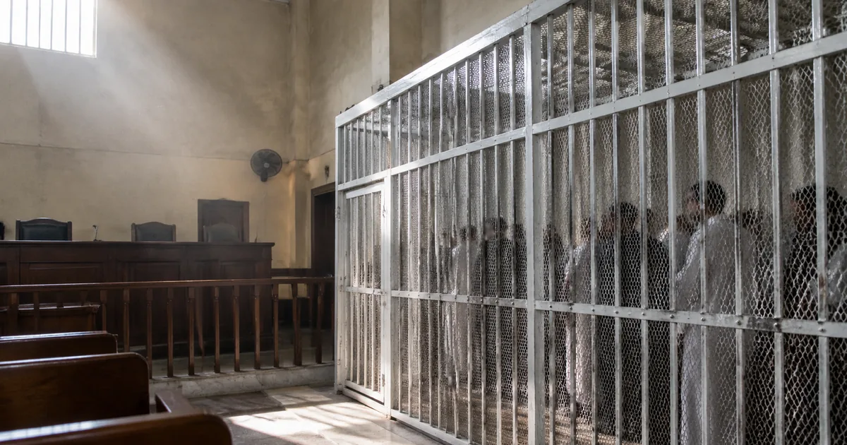 A tall white steel defendants cage stands in an Egyptian criminal courtroom, lit by daylight from a high window, with an empty judges bench to one side