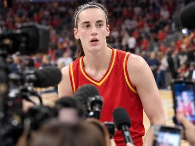 Caitlin Clark in an Indiana Fever uniform on the court, framed by a dense out-of-focus foreground of television cameras, microphones and raised phones