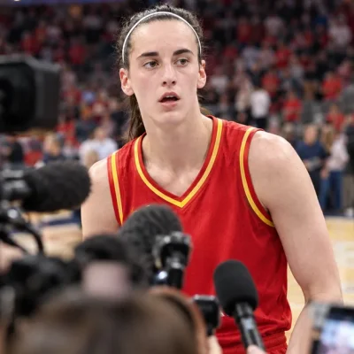 Caitlin Clark in an Indiana Fever uniform on the court, framed by a dense out-of-focus foreground of television cameras, microphones and raised phones