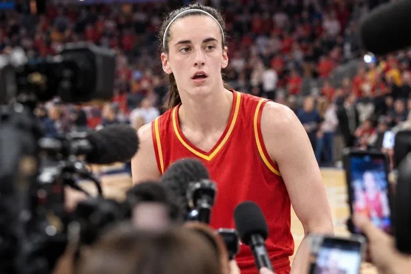 Caitlin Clark in an Indiana Fever uniform on the court, framed by a dense out-of-focus foreground of television cameras, microphones and raised phones