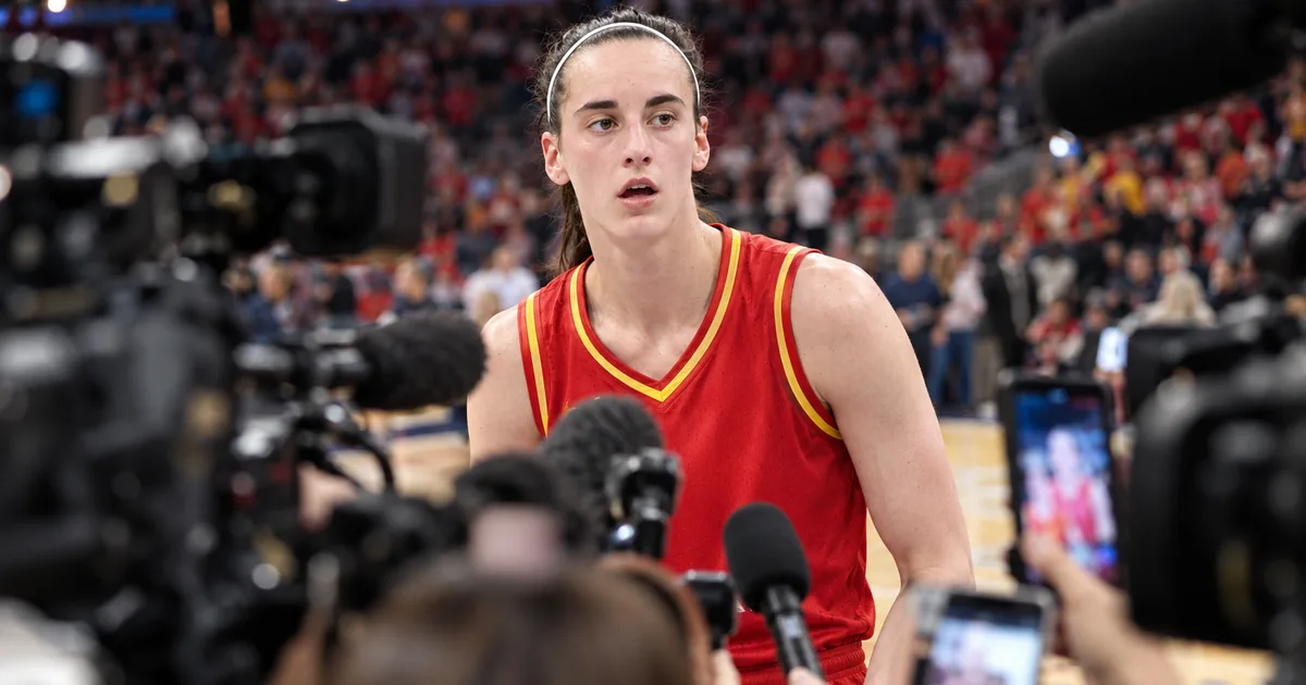 Caitlin Clark in an Indiana Fever uniform on the court, framed by a dense out-of-focus foreground of television cameras, microphones and raised phones