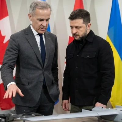 Mark Carney gestures toward a quadcopter and a fixed-wing drone on a display table as Volodymyr Zelenskyy looks on, with Canadian and Ukrainian flags behind them