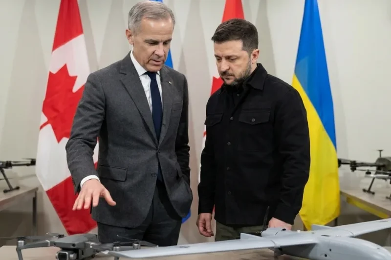 Mark Carney gestures toward a quadcopter and a fixed-wing drone on a display table as Volodymyr Zelenskyy looks on, with Canadian and Ukrainian flags behind them