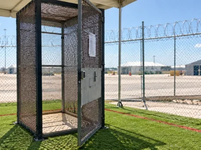 An empty phone-booth-sized steel mesh enclosure with a keypad-locked door standing on artificial turf beneath a tent canopy, with razor-wire-topped chain-link fencing and white detention tents behind it