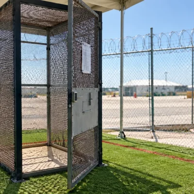 An empty phone-booth-sized steel mesh enclosure with a keypad-locked door standing on artificial turf beneath a tent canopy, with razor-wire-topped chain-link fencing and white detention tents behind it