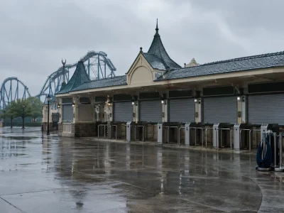 Empty amusement park ticket plaza on a wet overcast morning with closed ticket window shutters, metal turnstiles and a coiled queue rope