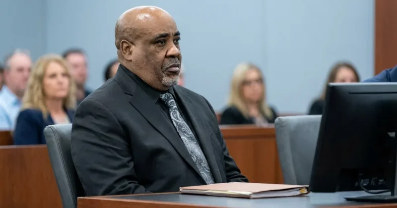 A bald man in his sixties with a grey goatee, wearing a dark suit and patterned grey tie, sits at a wooden courtroom defense table with a closed legal folder in front of him