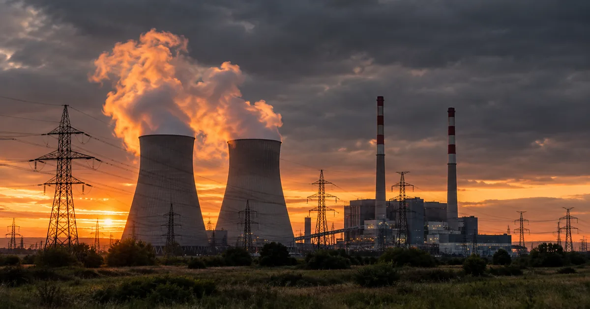 Cooling towers releasing steam plumes lit amber by a low sun behind a lattice of high-voltage transmission lines