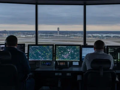 Two air traffic controllers seen from behind at a bank of radar and flight data screens inside a control tower, with runways and a distant tower visible through the windows at dusk