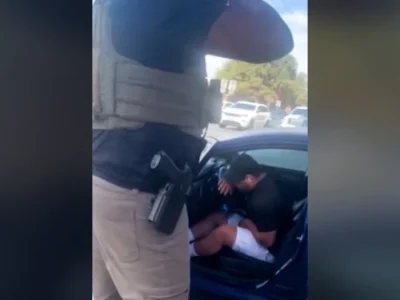 An armed federal agent in a tactical vest stands beside a blue car where Wilber Rafael Garces Perez sits hunched over after being shot, frame from video aired by CNN
