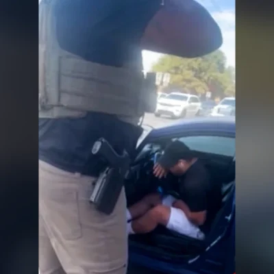 An armed federal agent in a tactical vest stands beside a blue car where Wilber Rafael Garces Perez sits hunched over after being shot, frame from video aired by CNN