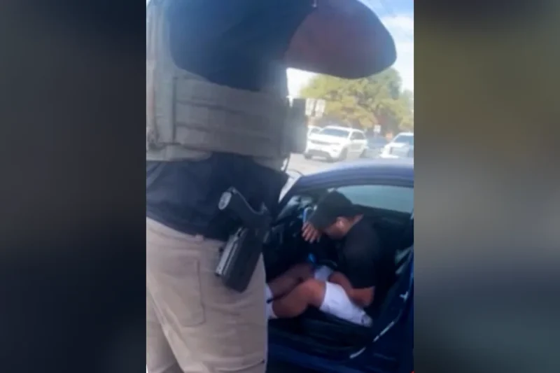 An armed federal agent in a tactical vest stands beside a blue car where Wilber Rafael Garces Perez sits hunched over after being shot, frame from video aired by CNN