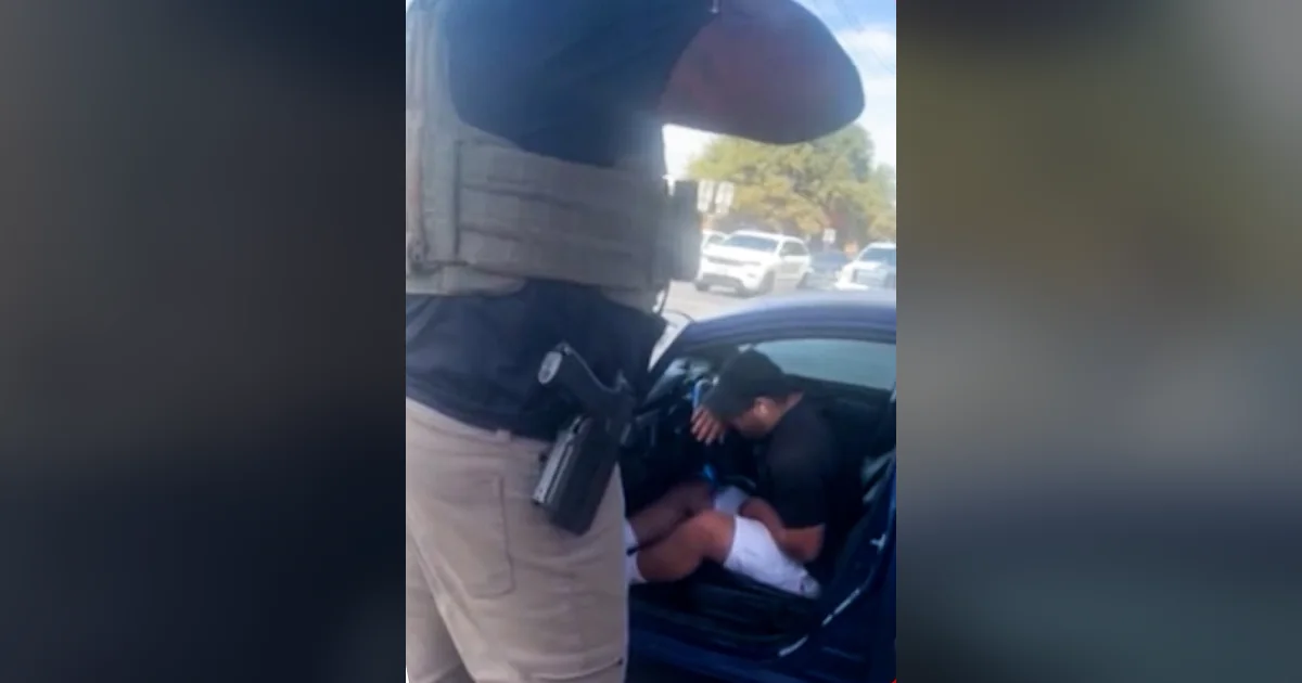 An armed federal agent in a tactical vest stands beside a blue car where Wilber Rafael Garces Perez sits hunched over after being shot, frame from video aired by CNN