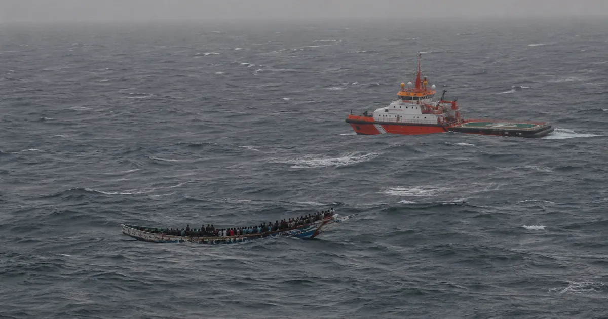Aerial view of a long wooden fishing boat crowded with people sitting low in heavy grey Atlantic swell, an orange and white maritime rescue vessel standing off nearby under an overcast sky