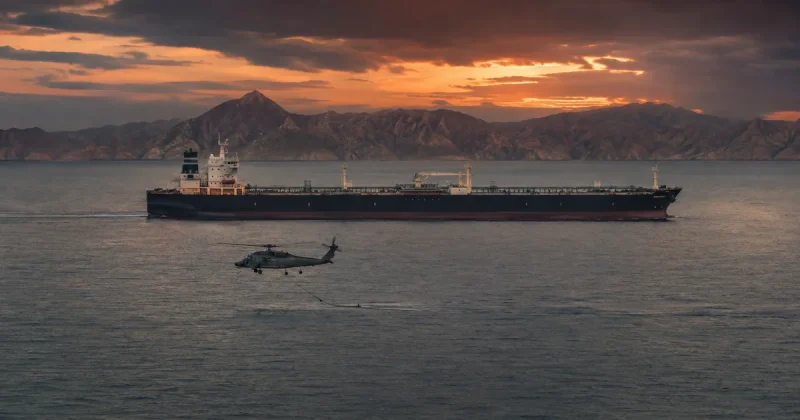 A laden crude oil tanker at dusk in the Strait of Hormuz as a military helicopter tows a minesweeping sonar sled nearby